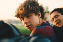 Close-up of a teenage boy sitting outside with friends, looking at the camera.